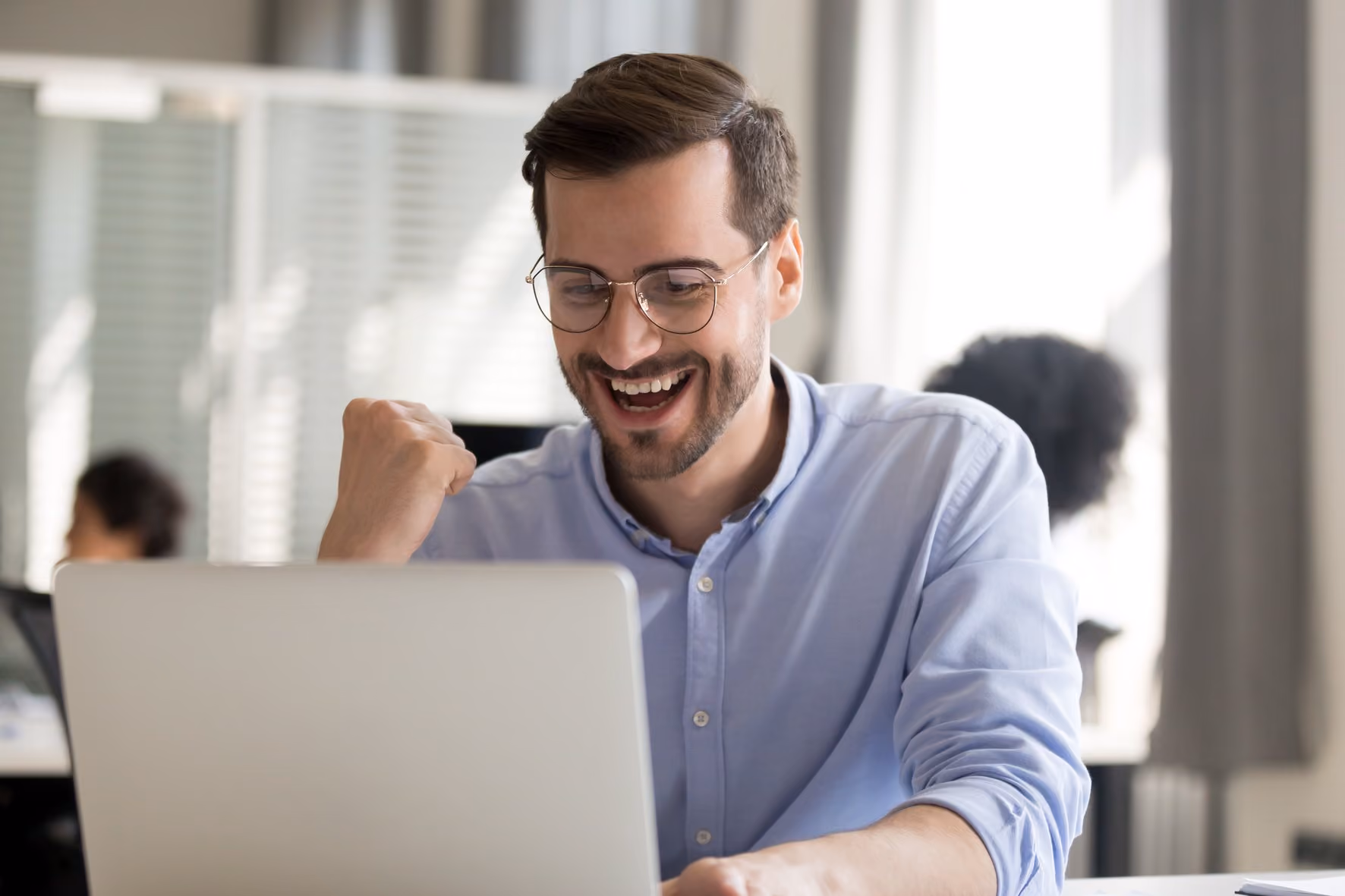 Man in glasses pumps his fist while cheering at a laptop in an open office.
