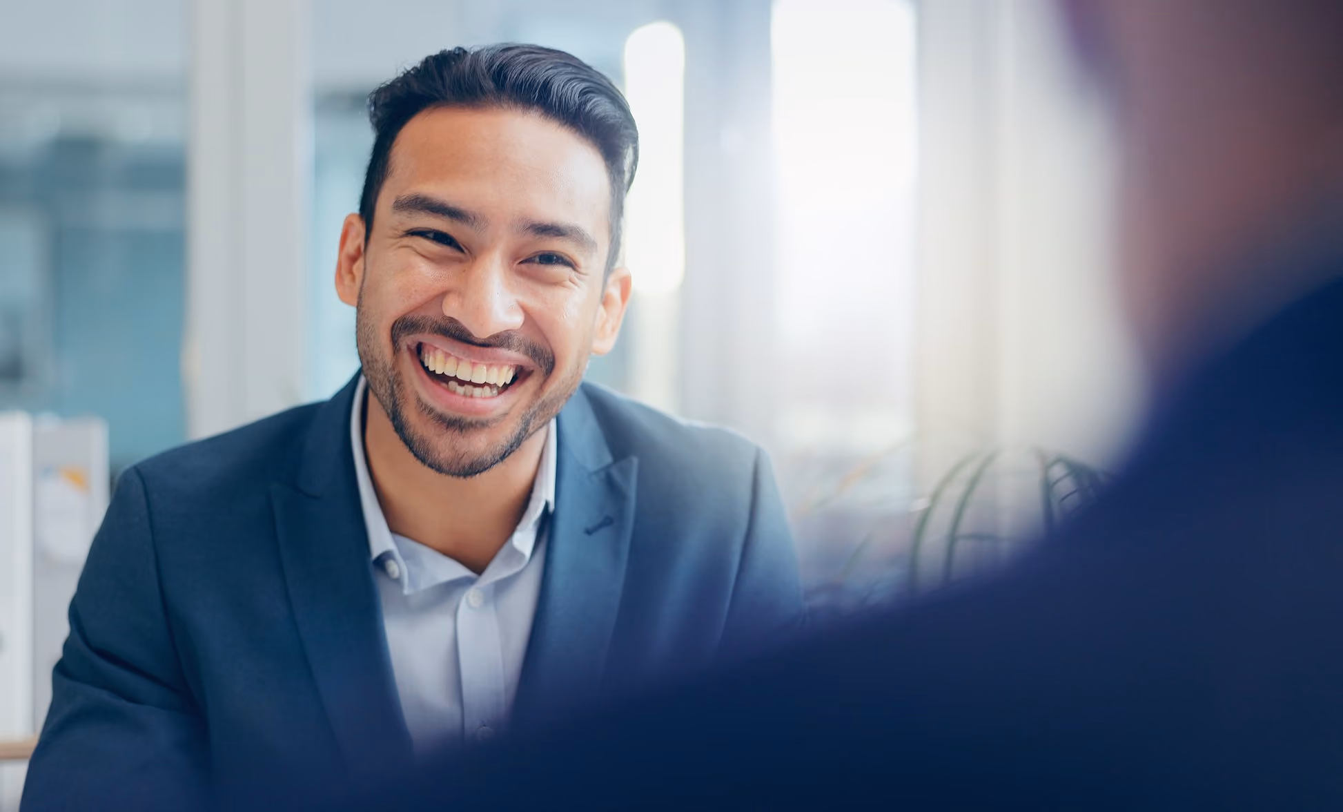 Laughing man in a navy suit talks with a colleague in a bright office.