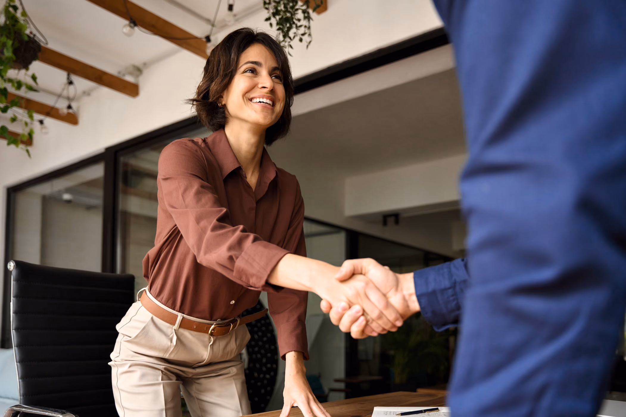 Smiling woman in a burgundy shirt shakes hands across a conference table.