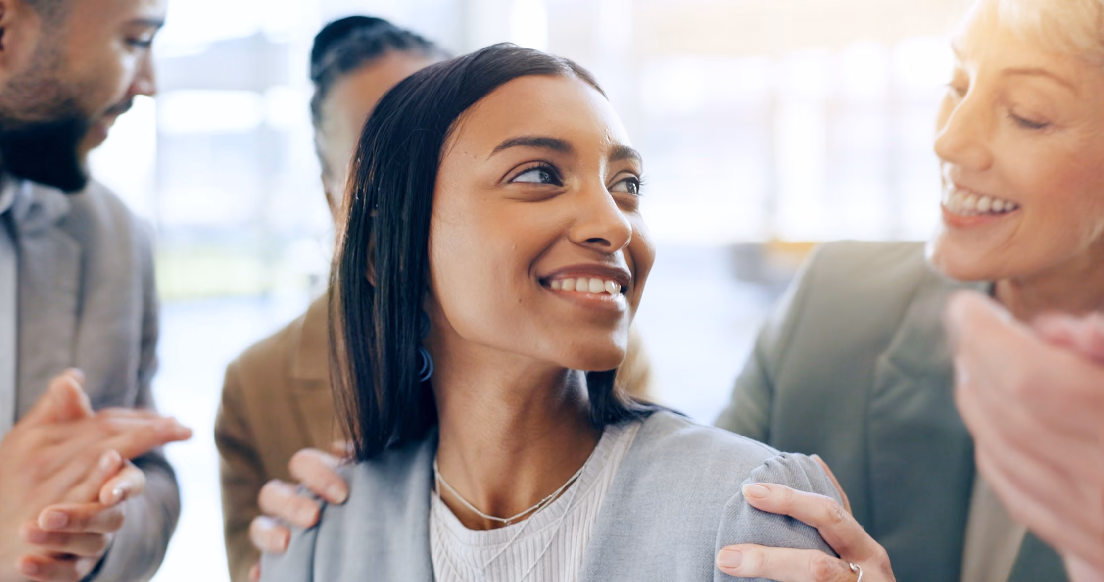 Smiling woman receives congratulatory shoulder touches from colleagues in an office.