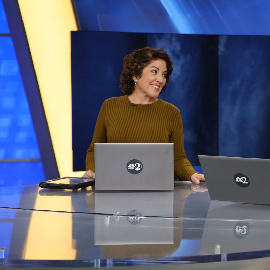 Anchor in a yellow sweater smiles at an NBC 2 news desk with laptops.