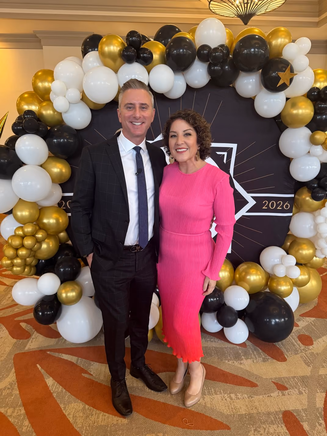 Couple in formal attire pose before a 2026 balloon arch at a gala.