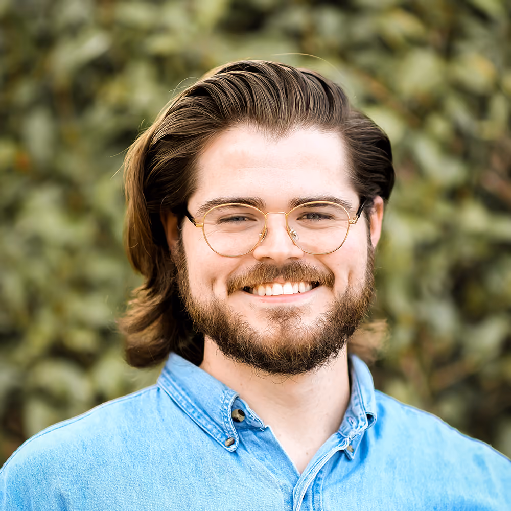 Smiling man with glasses and beard wearing a blue denim shirt in front of leafy green background.
