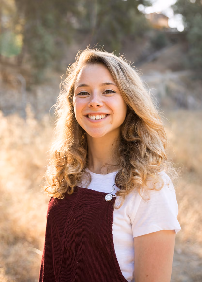 Smiling young woman with curly blonde hair wearing a white shirt and maroon overall outdoors.