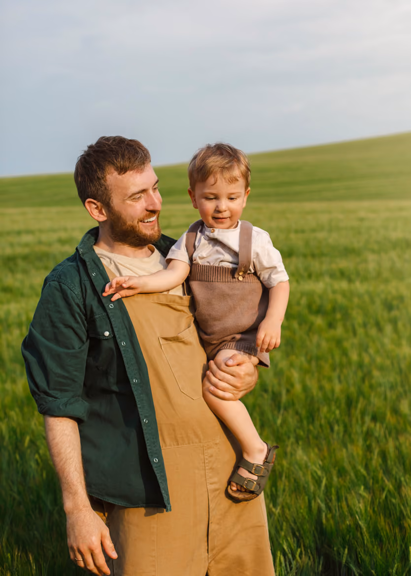 Smiling bearded man holding toddler in overalls in a green field under a blue sky.