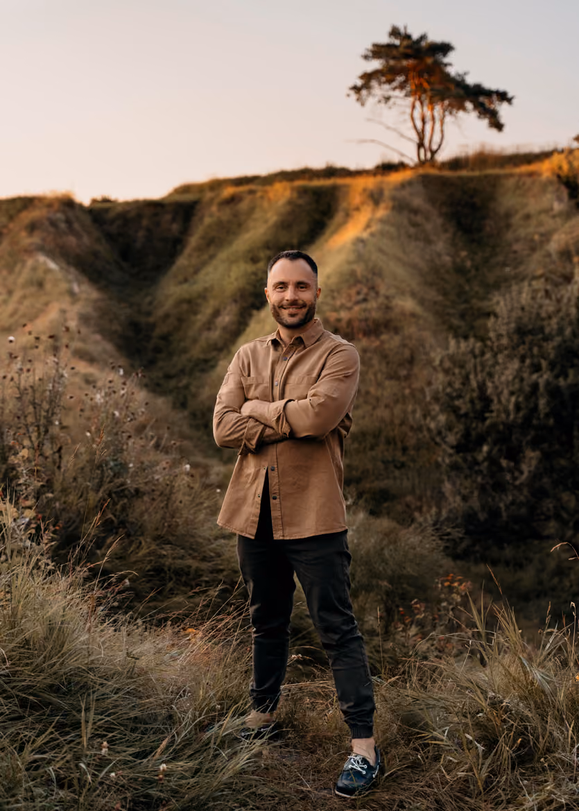 Man in brown shirt and black pants standing with arms crossed in grassy outdoor landscape at sunset.