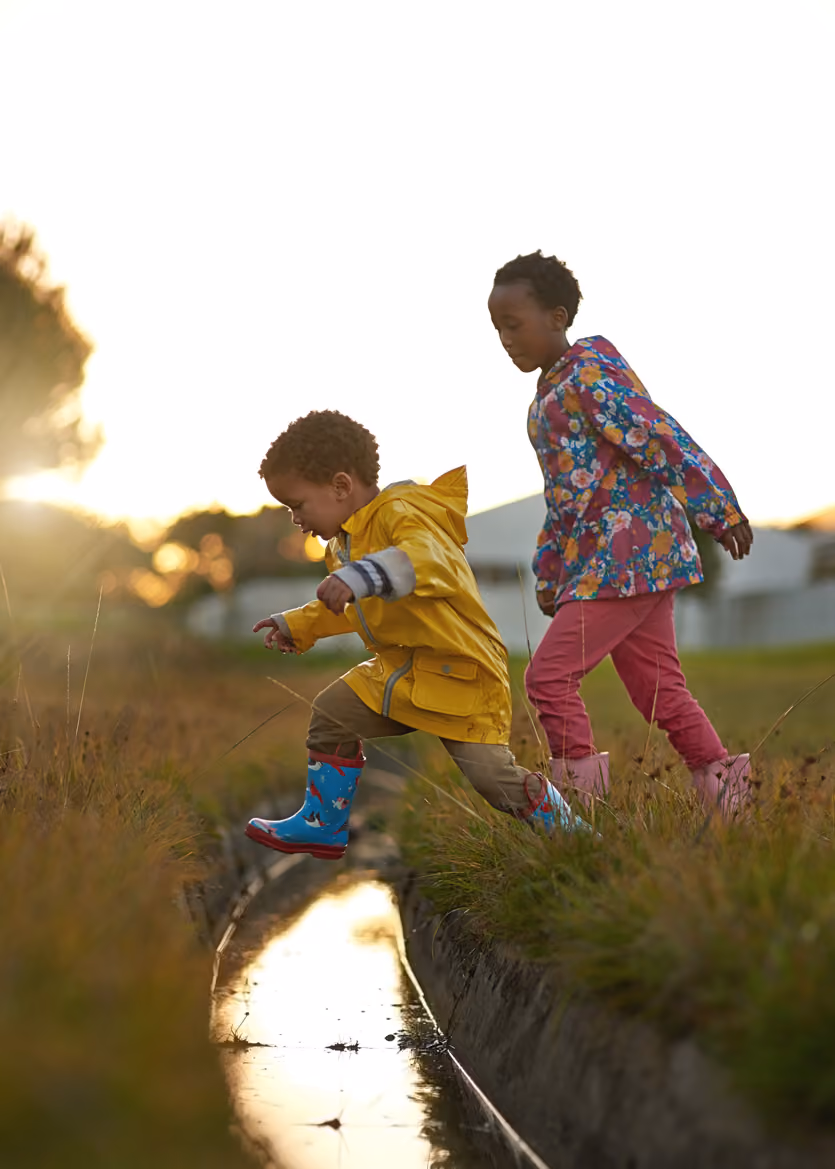 Young boy in yellow raincoat jumping over a narrow water ditch with girl in floral jacket behind him.