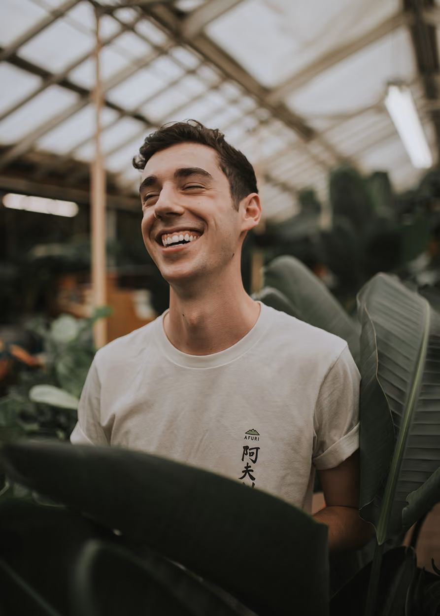 Smiling young man in a white t-shirt stands among large green leaves inside a glasshouse.