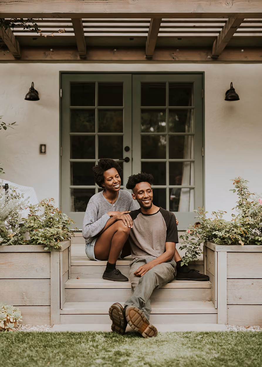 Couple sitting on wooden steps outside a house with green doors and plants on either side.