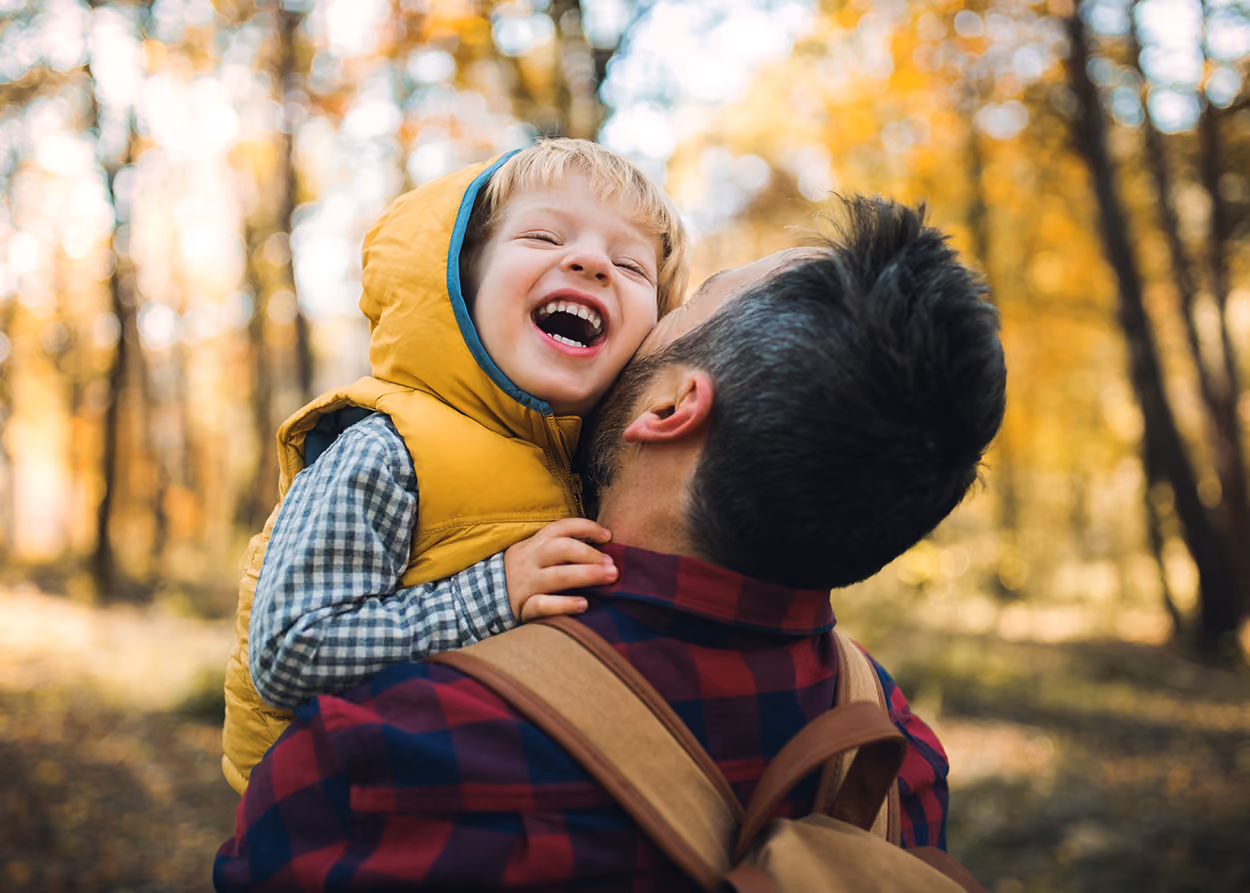 A loving father holding his laughing toddler son in a sunlit autumn forest, illustrating the secure, supportive connection that helps children grow and learn.