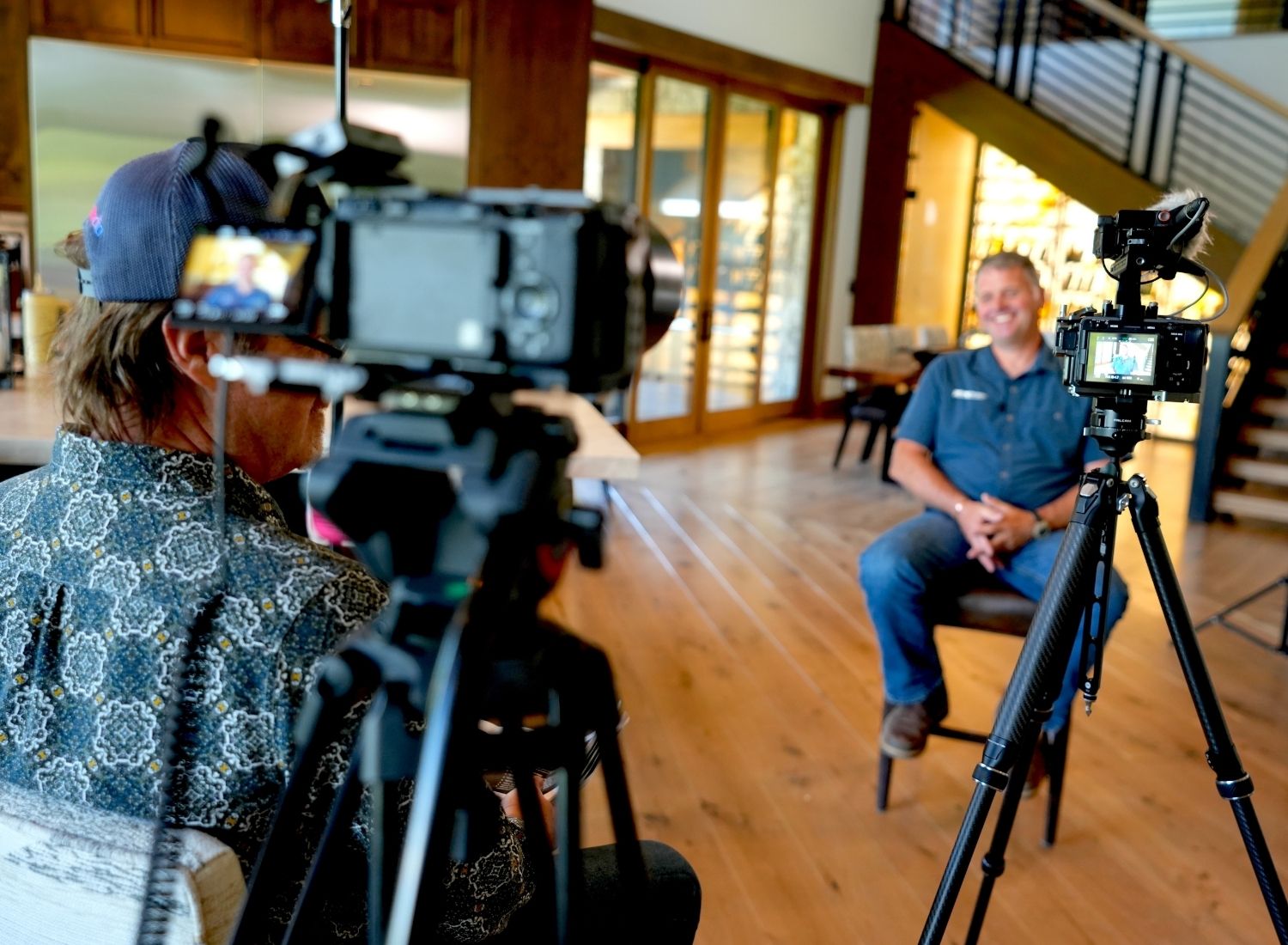 Man in a patterned shirt filming an interview with another man sitting in a chair smiling.