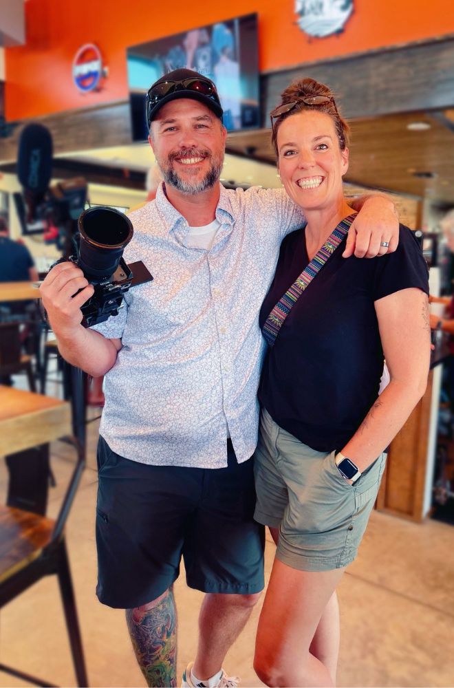 Smiling man holding a camera with arm around a smiling woman in casual attire inside a restaurant.