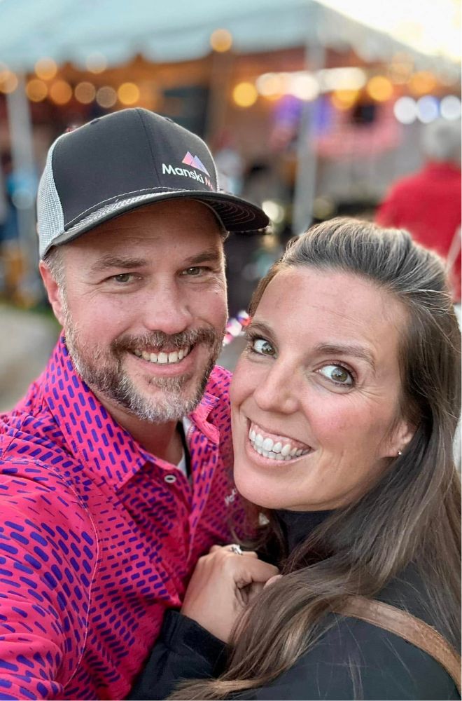 Smiling man in a cap and pink shirt takes a close selfie with a woman with long hair and bright eyes.