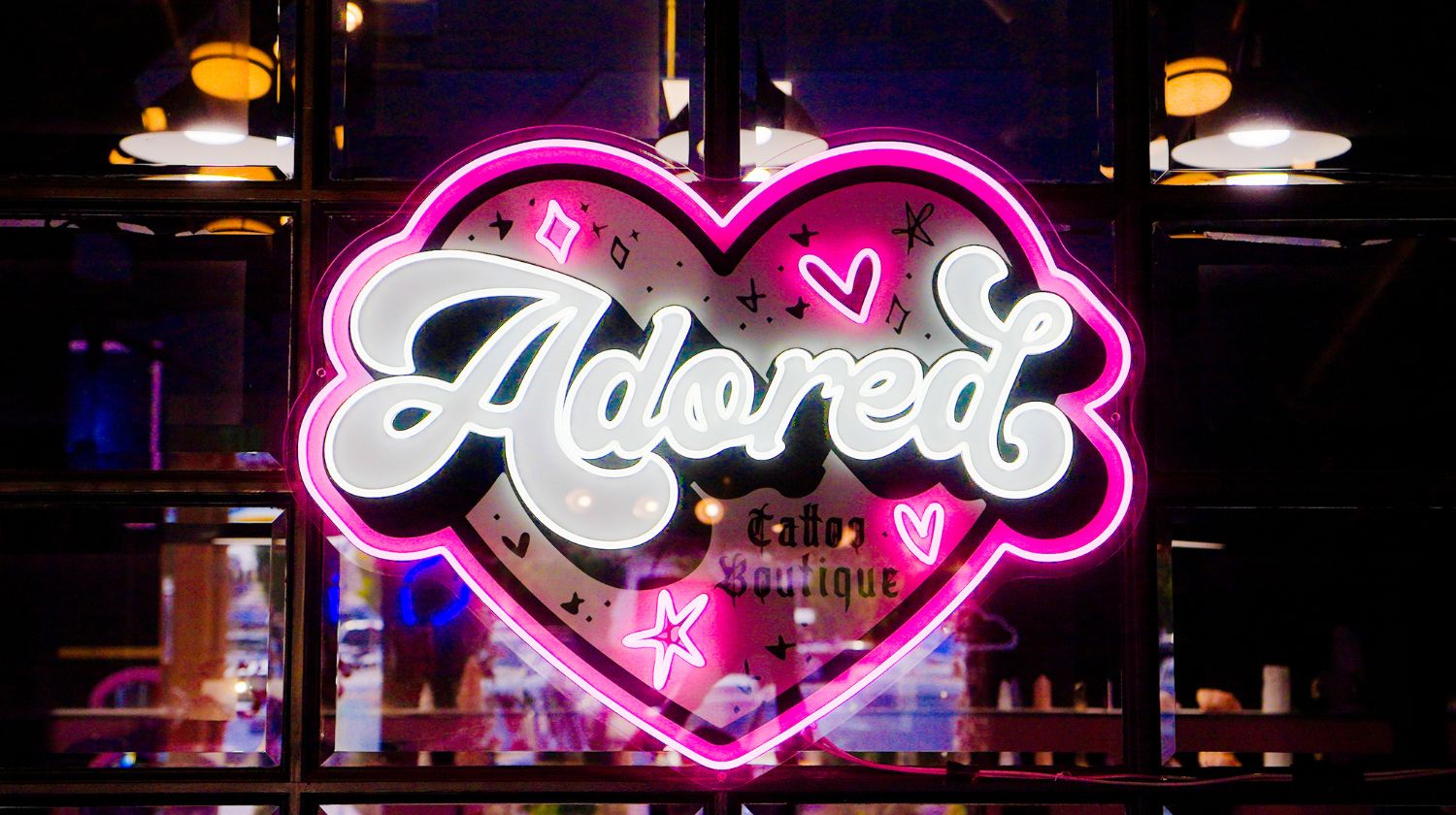 Neon sign in a heart shape with the word 'Adored' and decorations in pink and white.