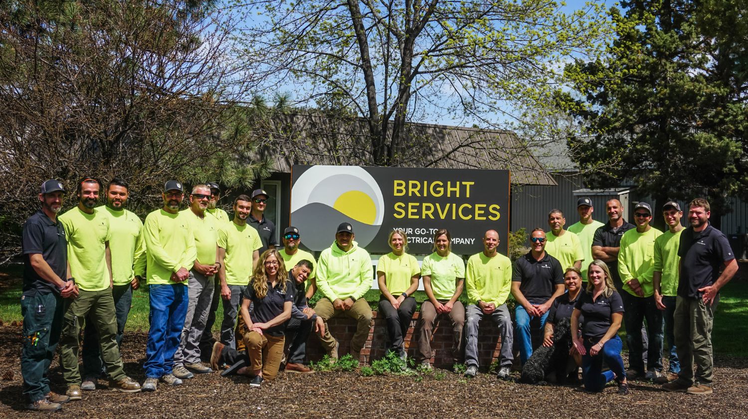 Group of people posing outside near a sign that reads Bright Services, an electrical company.