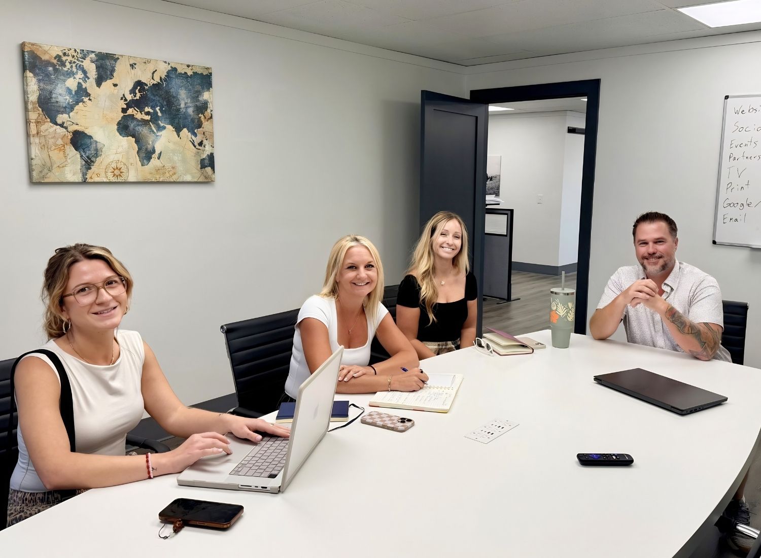 Four people smiling in a meeting room with laptops, notebooks, and a wall map behind them.