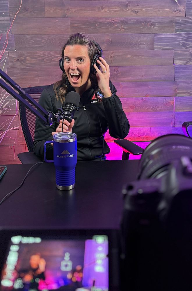 Woman wearing headphones speaking into a microphone with a blue Manski Media cup on the desk.