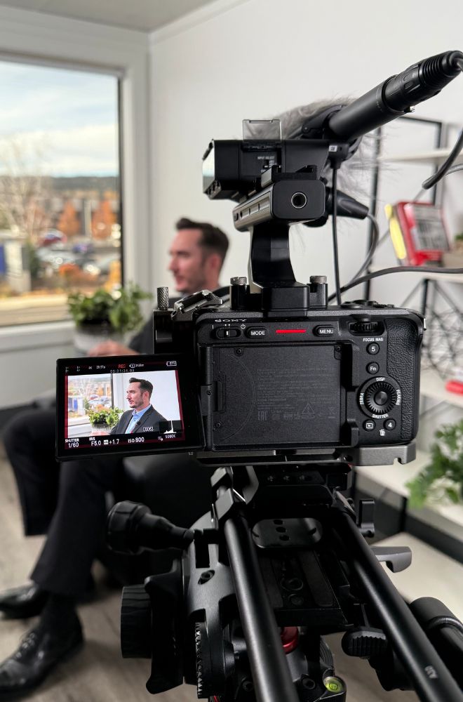 Digital camera filming a man in a suit during an indoor interview in a bright room with a window and shelves.