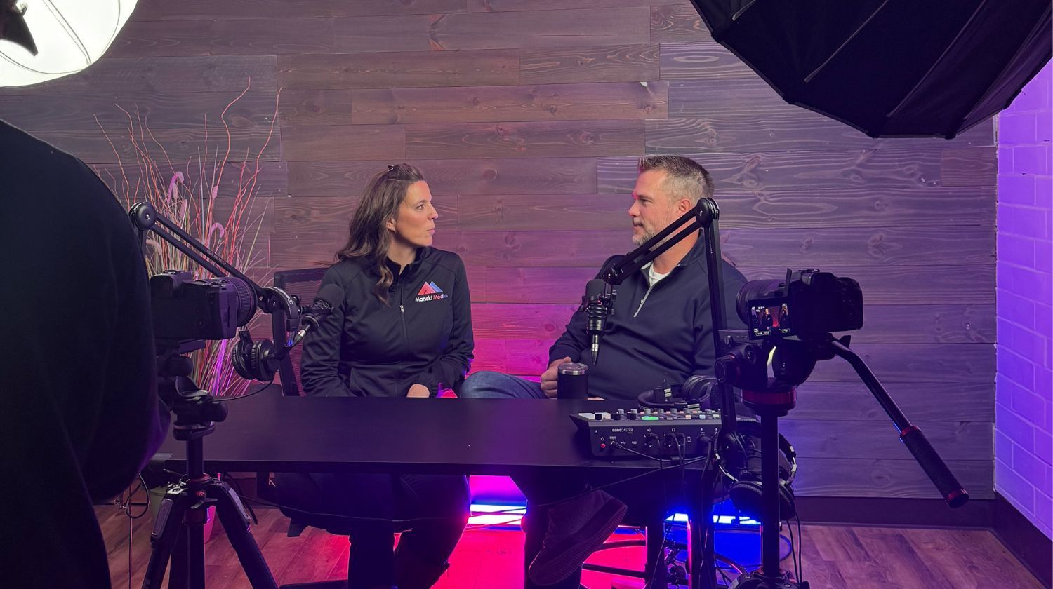 Two people seated at a podcast setup with microphones, cameras, and studio lighting in a wood-paneled room.
