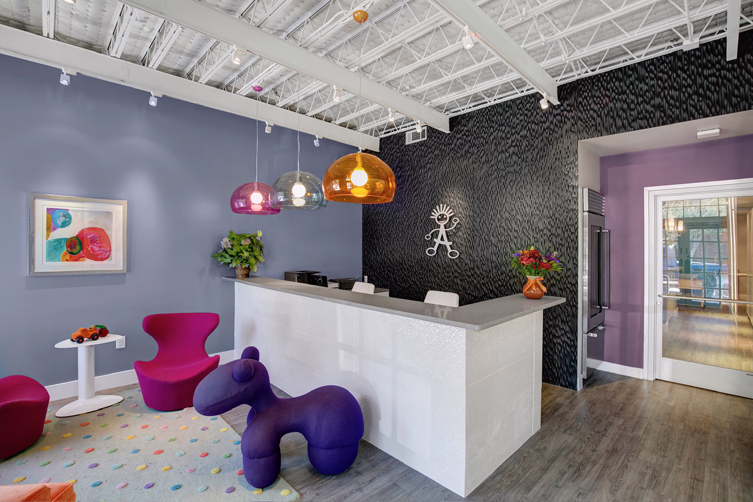 Modern reception area with a white front desk against a textured black wall featuring a decorative metal wall art, three colorful pendant lamps overhead, and a seating area with vibrant purple and magenta chairs and a playful purple animal-shaped seat on a light carpet with multicolored dots.
