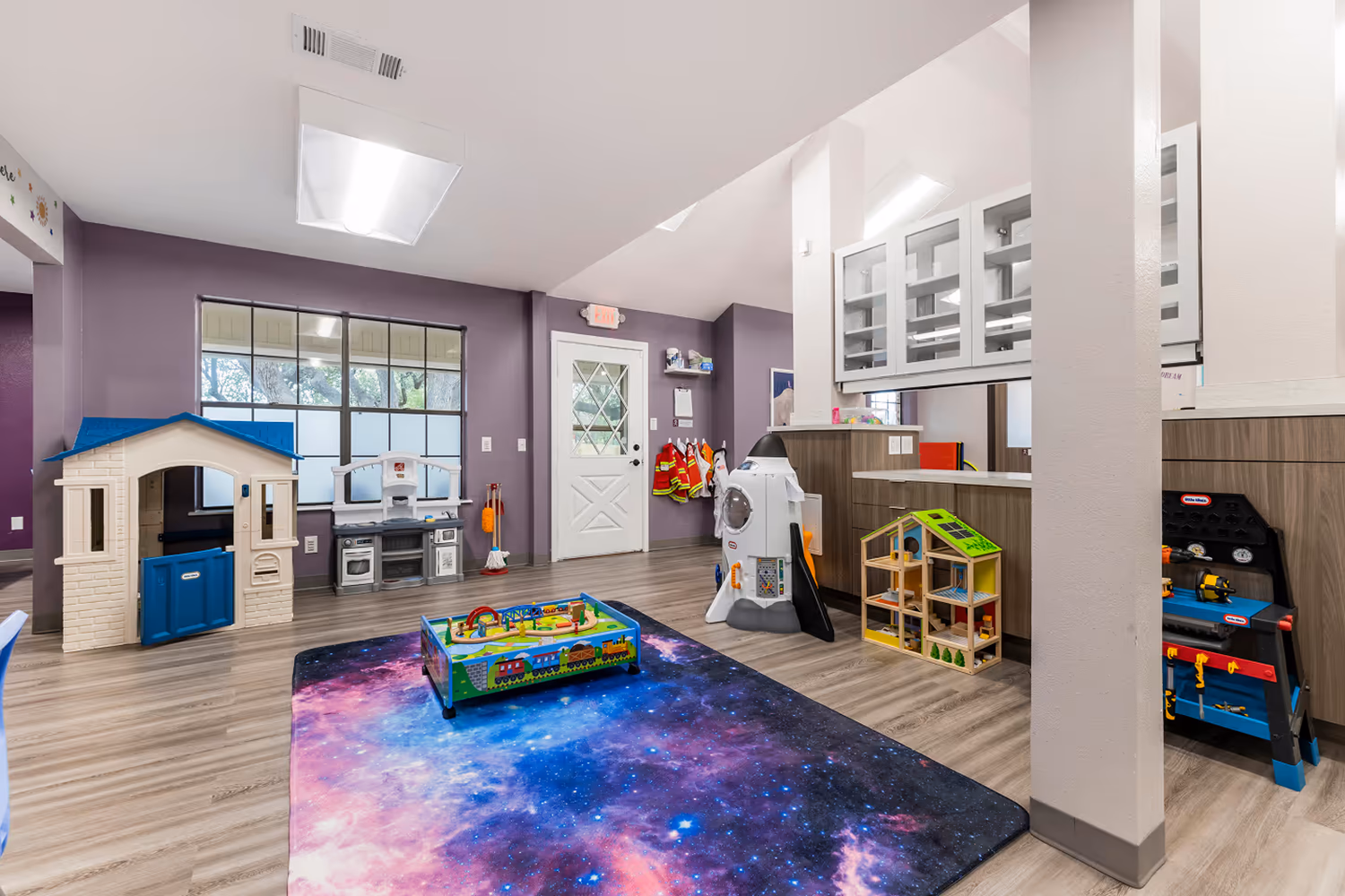 Bright indoor children's playroom with light wood flooring and vibrant purple walls, featuring a galaxy-themed rug, a white and blue playhouse, toy kitchen set, rocket-shaped toy, dollhouse, and various other playsets near a white door and large windows.