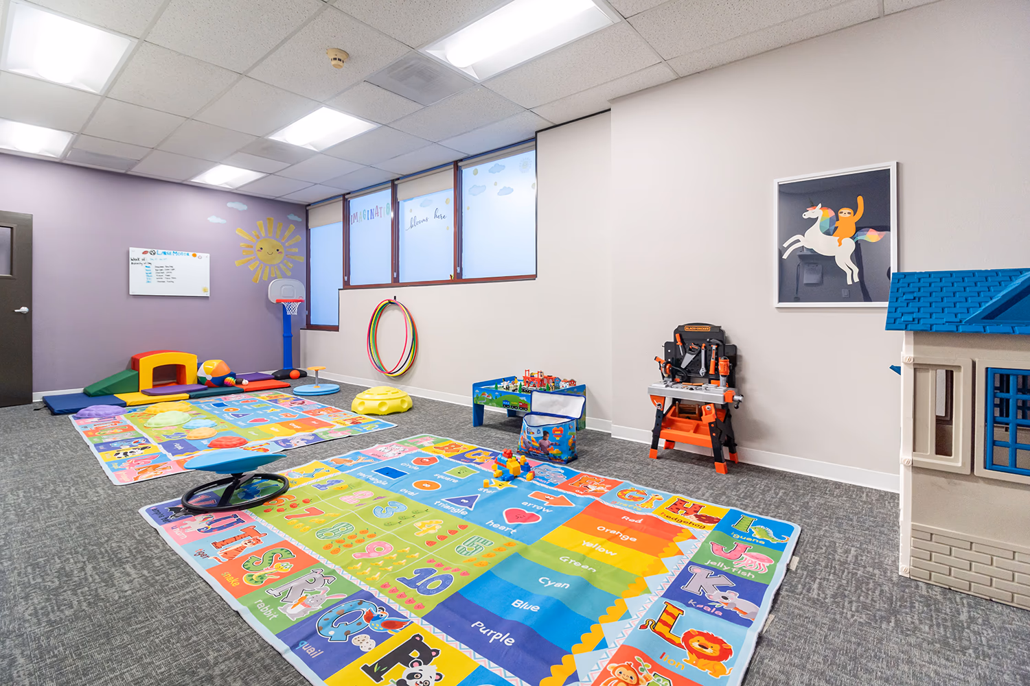 Bright kids' playroom with colorful educational rugs featuring numbers, letters, and colors, a small basketball hoop, toy house, play tools, hula hoops, and a wall painting of a unicorn and girl.