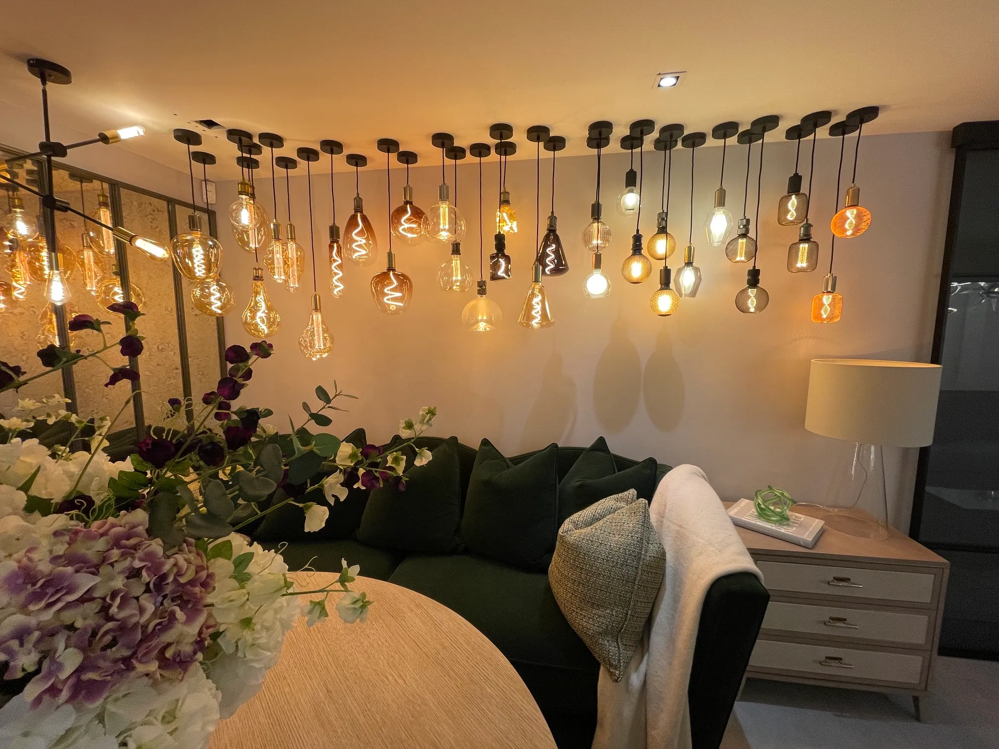 Cozy living room corner featuring a green velvet sofa with cushions and a white throw, a wooden side table with a lamp and decorative items, a round wooden table in the foreground with a bouquet of white and purple flowers, and a ceiling installation of various vintage-style hanging Edison bulbs illuminating the space warmly.