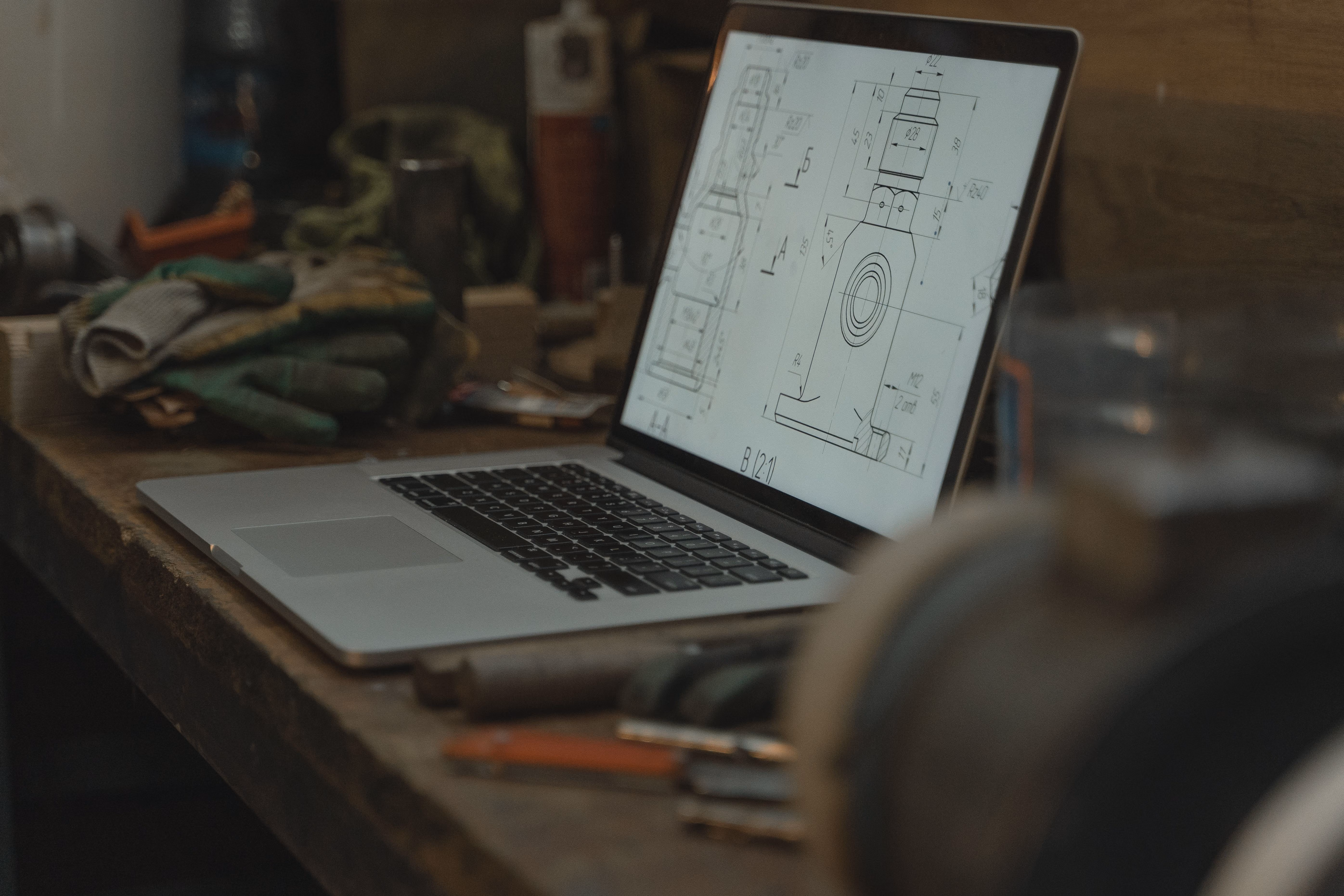 Laptop on a wooden workbench displaying a technical drawing or blueprint of a mechanical part, with tools and work gloves nearby.