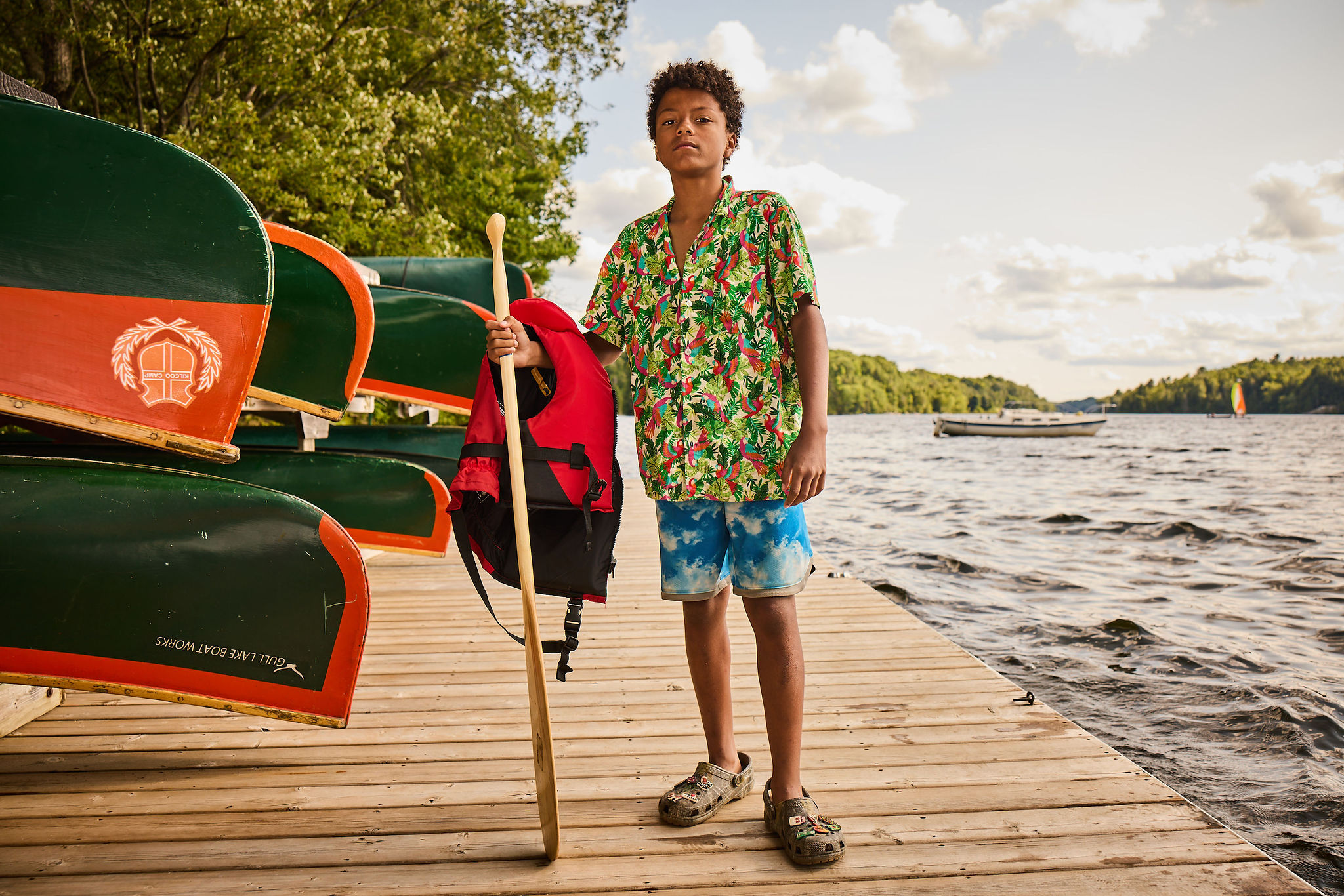 A camper holding a paddle and life jacket on the dock, forest-green canoes with orange crest badges stacked beside him, Gull Lake behind