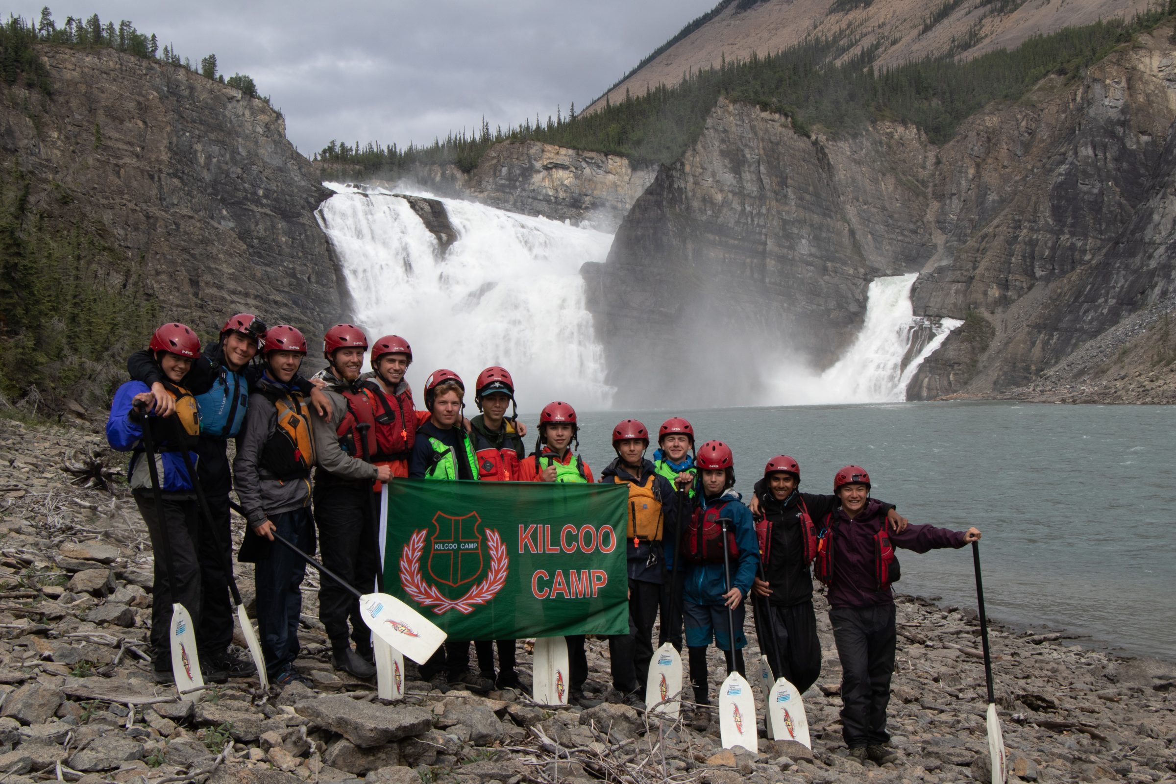 A group of Kilcoo campers and staff posed together at a waterfall on a canoe trip, holding a Kilcoo Camp banner