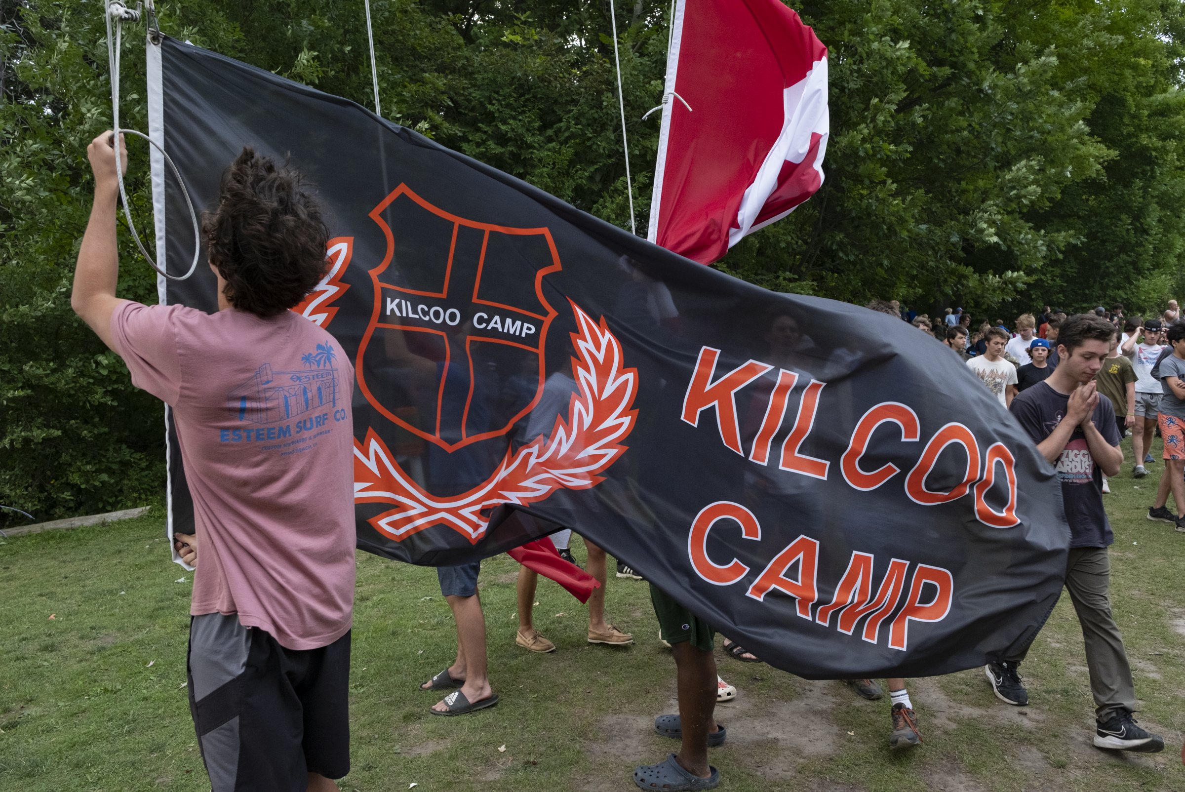 Campers carry the large black and orange Kilcoo Camp banner across the field during flag raising, with a Canadian flag and the whole camp gathered behind.