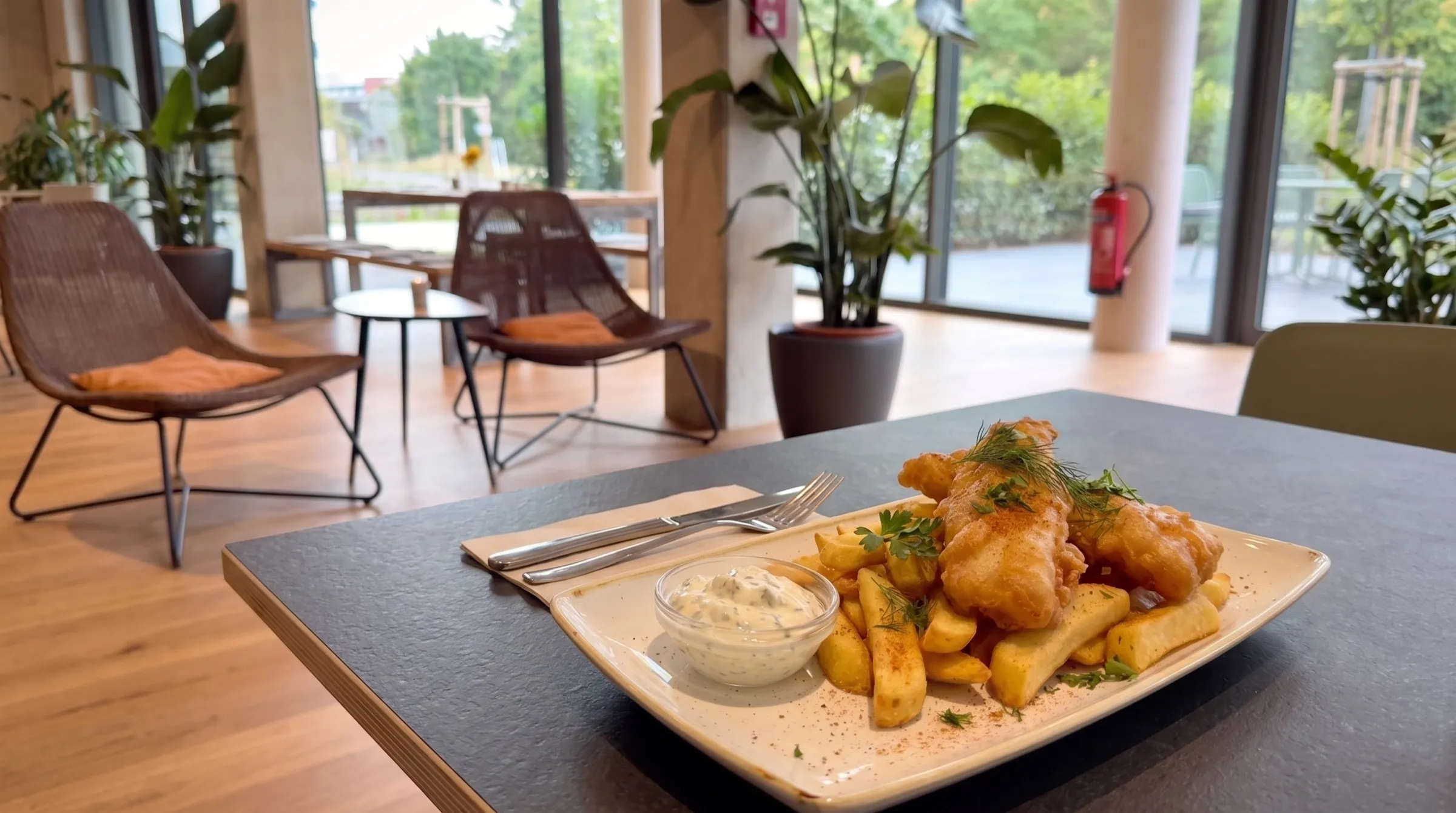 Teller mit Fisch und Fresh Fries auf einem Tisch im THE COURT, dahinter die Lounge-Ecke mit Korbsesseln und Fensterfront