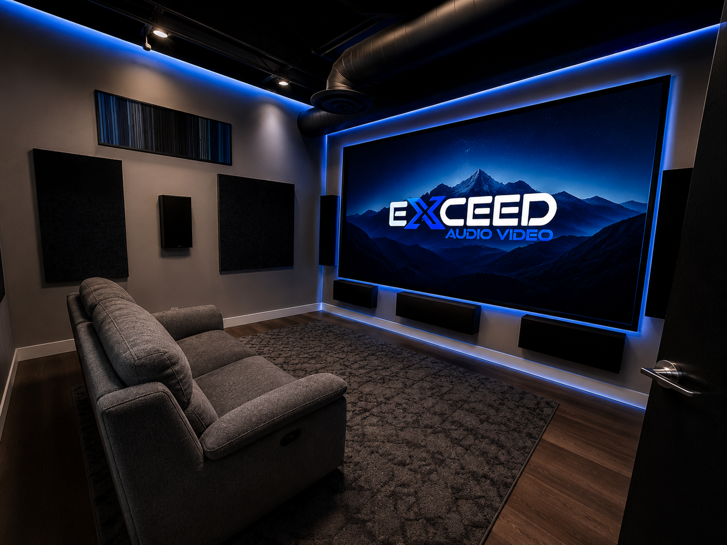 Modern home theater room with gray couch, dark rug, wall speakers, and large screen showing Exceed Audio Video logo.