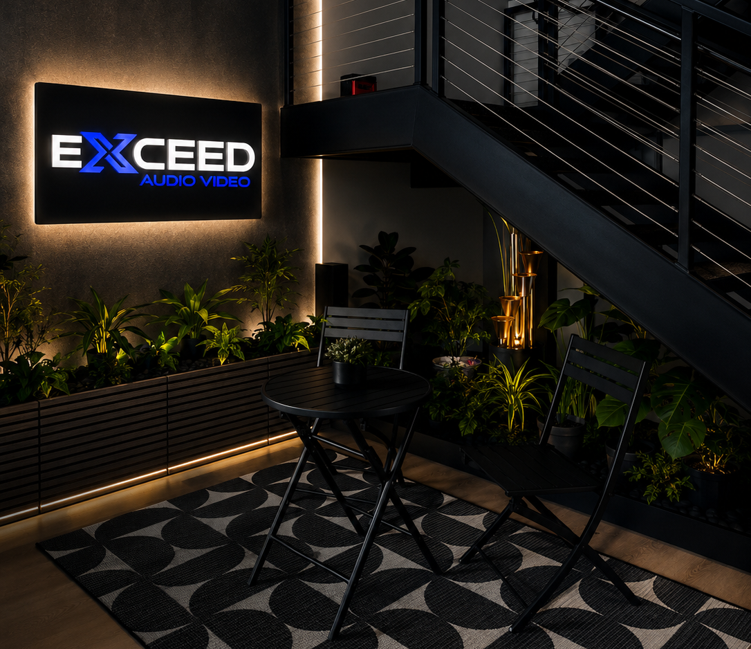 Modern seating area with black table and chairs, plants, and illuminated Exceed Audio Video sign on wall.
