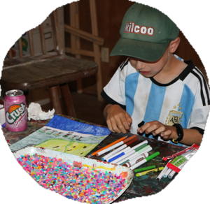 A young camper in a green Kilcoo cap works on a craft at a table covered in markers and beads.