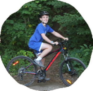 A camper in a helmet rides a mountain bike along a dirt trail through dense green forest.