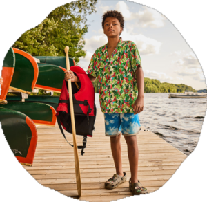 A camper stands on a lakeside dock holding a paddle and a red life jacket beside stacked green canoes.