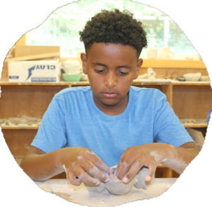A camper shapes a lump of wet clay with both hands at a worktable in the pottery studio.