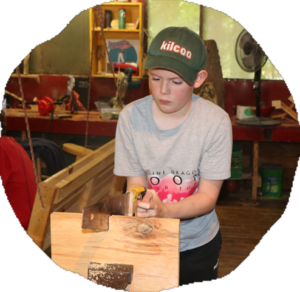 A camper in a green Kilcoo cap shapes a block of wood with a hand tool at the camp handicraft workbench.