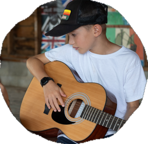 A camper in a black ball cap plays an acoustic guitar inside a sunlit camp building.