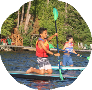 Two campers in life jackets kneel on stand up paddleboards, paddling past the camp dock and shoreline.