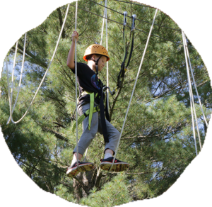 A camper in an orange helmet and safety harness balances on a high ropes course element among tall pines.