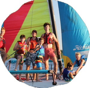 Campers in life jackets crowd the deck of a catamaran under a rainbow striped sail.