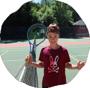 A smiling camper stands on an outdoor tennis court holding a racket with arms spread wide.