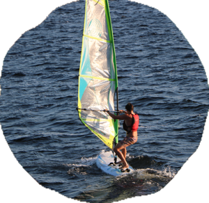 A camper in a red life jacket rides a windsurfer with a yellow and white sail across open lake water.