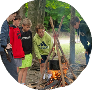 Five campers gather around a small cooking fire with a pot hung from a tripod of branches in the woods.