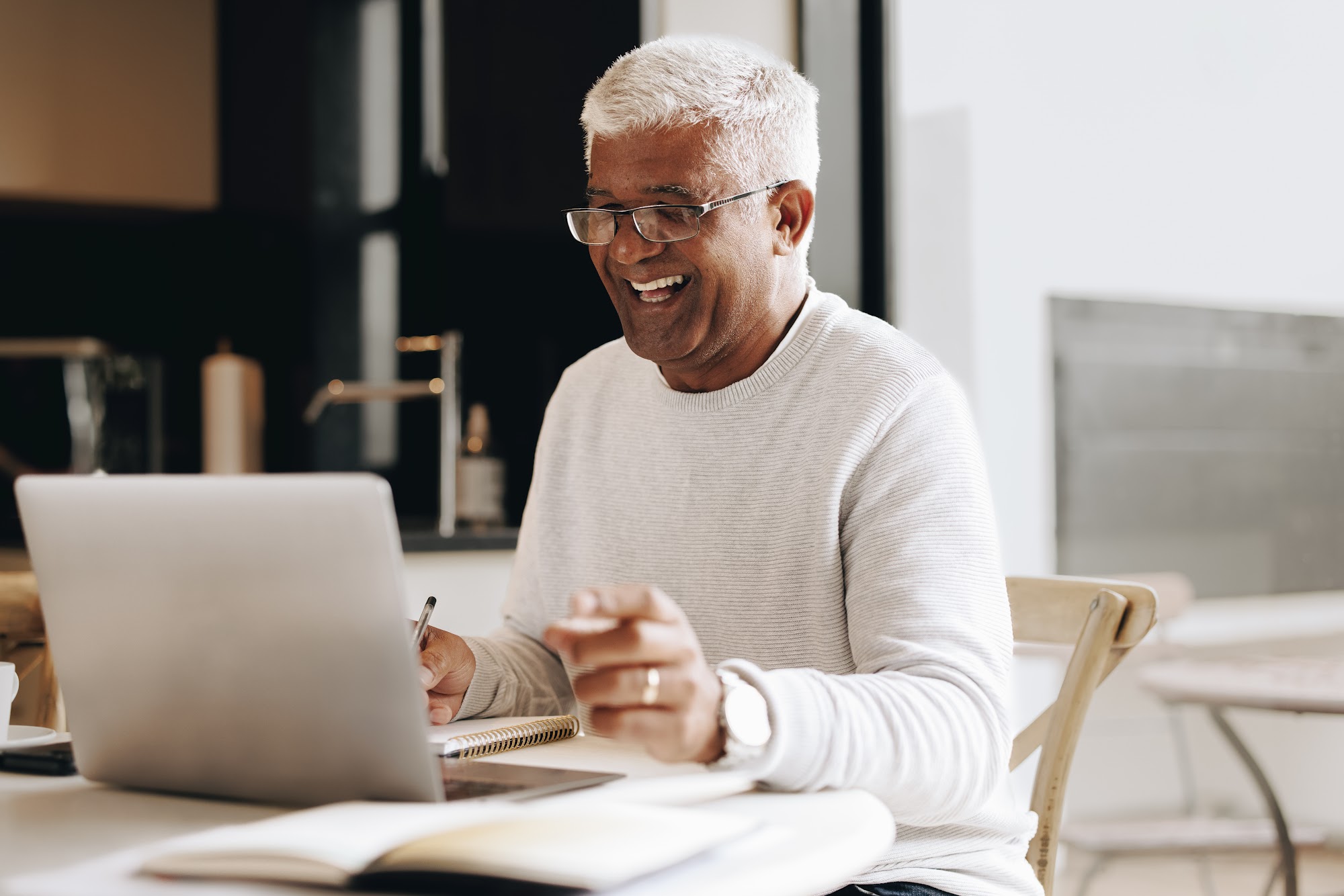 A silver-haired man wearing glasses and a cream sweater laughs while taking notes beside his laptop at a kitchen table.