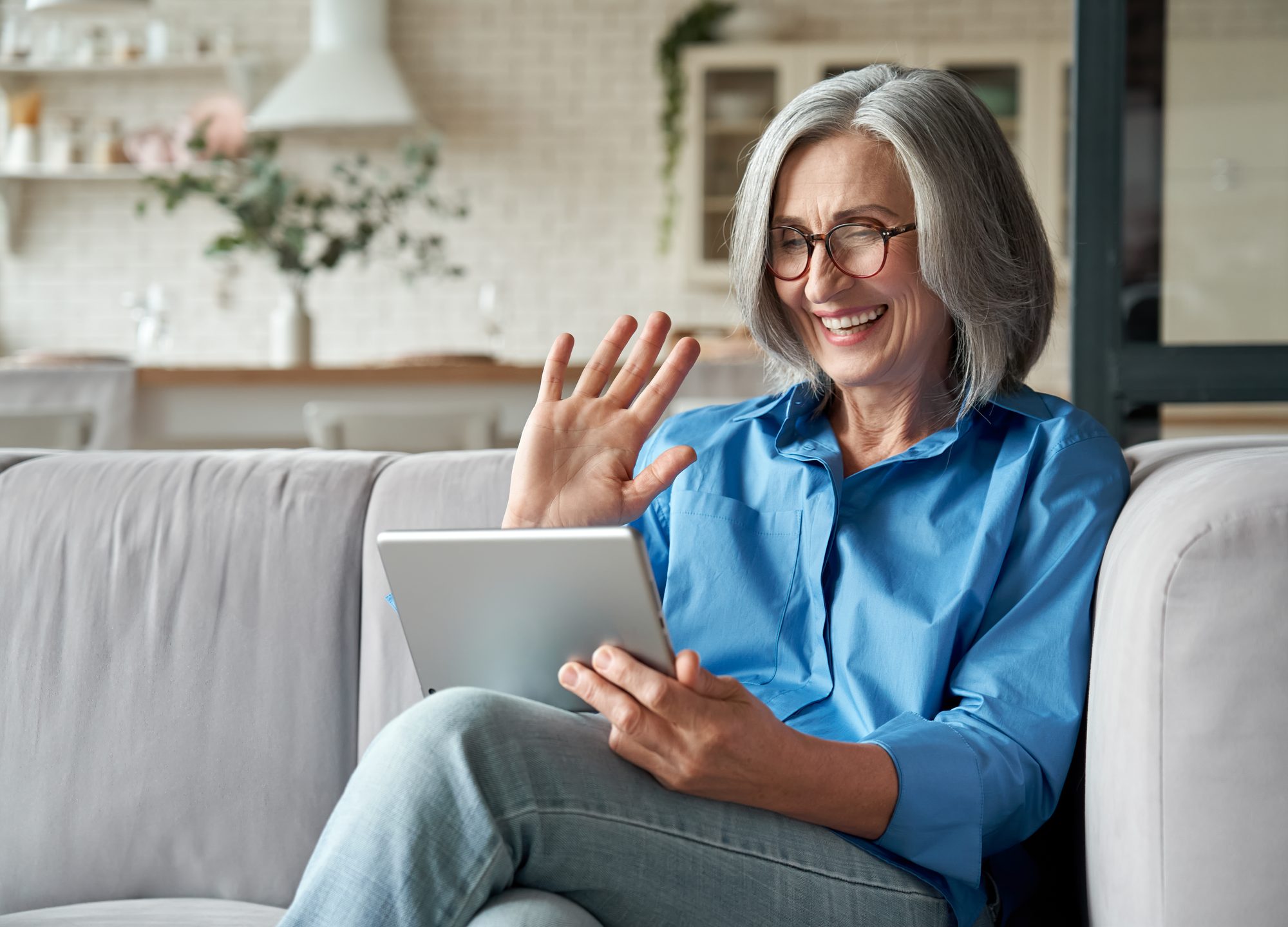 An older woman smiling and waving while holding a tablet during a video call at home.
