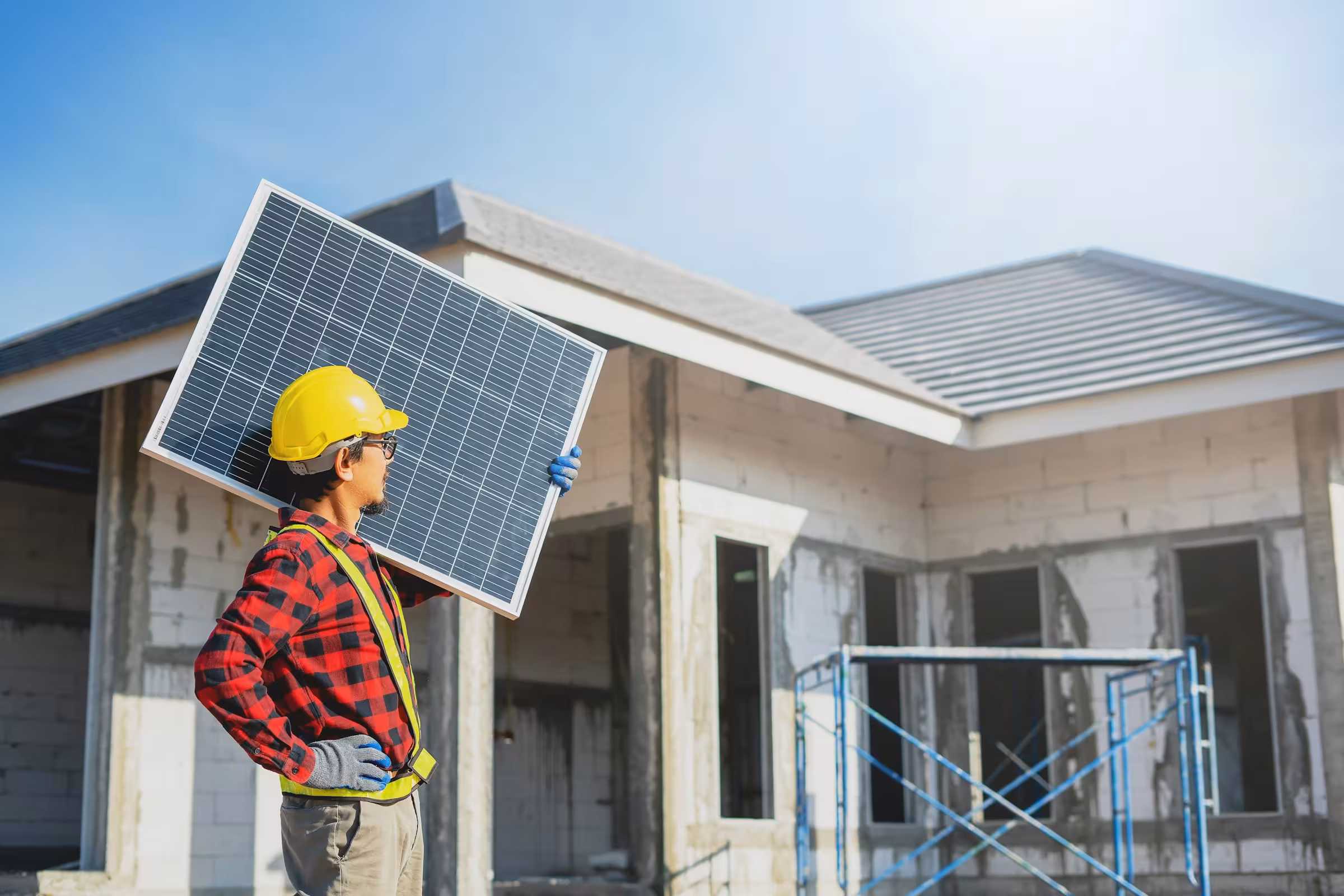 Construction worker wearing a yellow helmet and red plaid shirt carrying a solar panel at a building site.