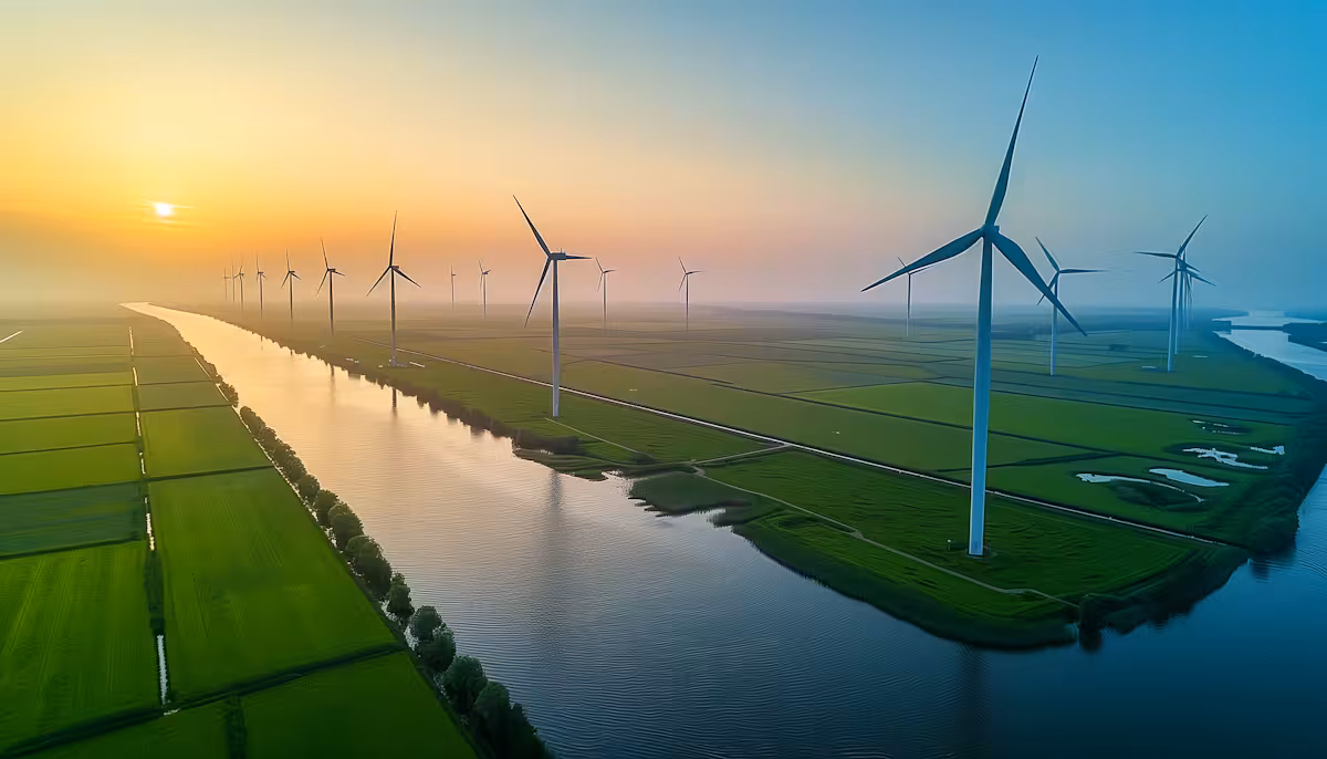 Wind turbines along a river at sunrise over green farmland under a clear sky.